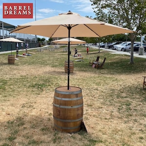 May include: A tan umbrella with a black frame is over a wooden barrel with a brown top. The barrel is on a grassy field with other barrels and people playing cornhole.