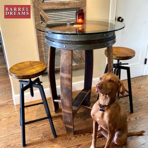 May include: A brown and black dog sits in front of a wooden table with a glass top. The table is made from a reclaimed wine barrel and has two black metal stools. The table is labeled "Barrel Dreams".