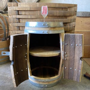 May include: A repurposed wooden barrel cabinet with two doors and a shelf inside. The cabinet is made from a wine barrel, with metal bands and wooden staves. A wine glass with the text "Barrel Dreams" sits on top.