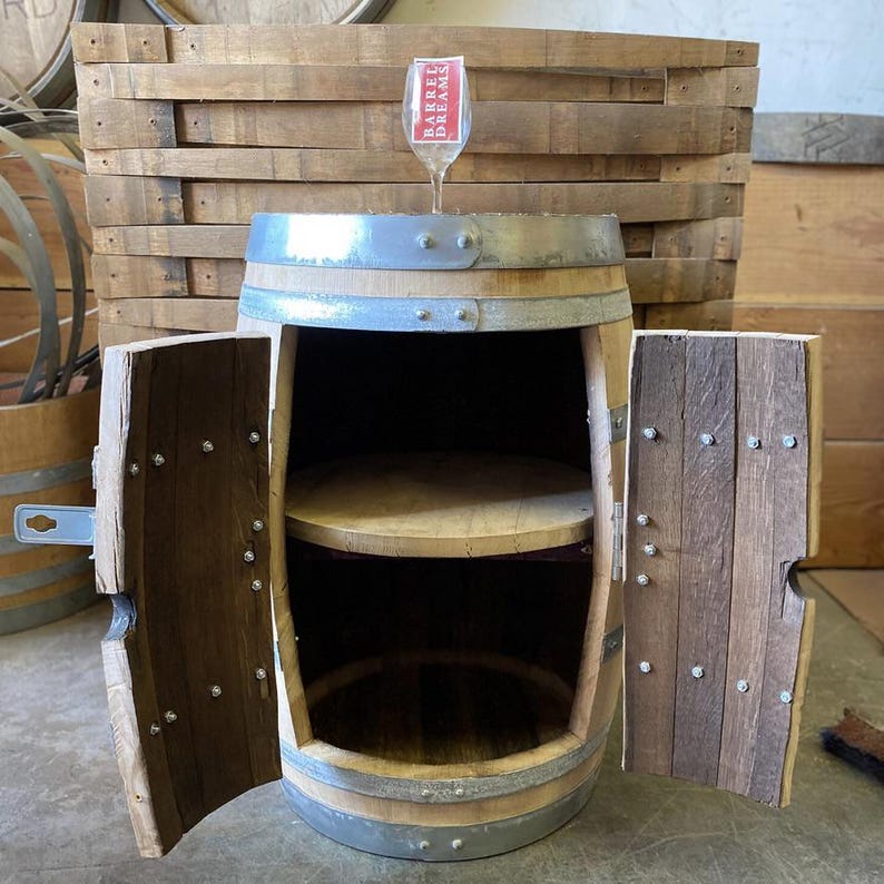 May include: A repurposed wooden barrel cabinet with two doors and a shelf inside. The cabinet is made from a wine barrel, with metal bands and wooden staves. A wine glass with the text "Barrel Dreams" sits on top.