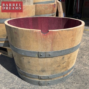 May include: A wooden barrel planter with metal bands. The inside of the barrel is painted a deep burgundy red. The barrel is sitting on a wooden pallet. The text "BARREL DREAMS" is visible on the barrel.