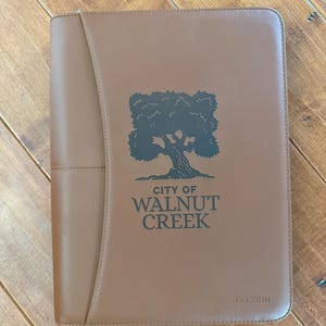 May include: A brown leather portfolio with the text "CITY OF WALNUT CREEK" and a tree graphic. The portfolio has a stitched design and the brand name "COSSINI" is printed on the bottom right corner.