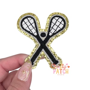 May include: Embroidered patch featuring two crossed lacrosse sticks in black and white, set against a gold glitter background. The patch has a white border and a black outline. The patch has the text "Crafty Patch Studio".