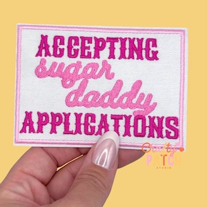 May include: A white rectangular patch with pink embroidered text that reads "ACCEPTING sugar daddy APPLICATIONS". The patch has a pink border and is held by a hand against a yellow background.