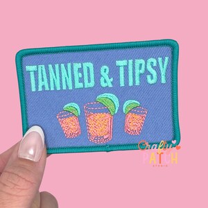 May include: Embroidered patch with the words "TANNED & TIPSY" in turquoise on a blue background. The patch features three cocktail glasses with orange liquid and lime wedges. The patch has a turquoise border and is held by a hand.