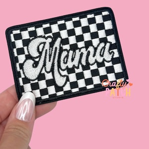May include: A rectangular patch with a black and white checkered pattern. The word "Mama" is embroidered in white, cursive lettering. The patch has a black border and the text "Crafty Patch Studio" is visible.