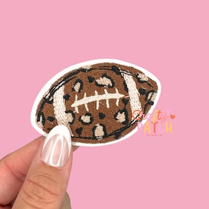 May include: An embroidered patch in the shape of a football with a leopard print design. The patch is brown with black spots and white stitching. The patch is held by a person's hand against a pink background. The text "Crafty Patch Studio" is visible.