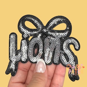 May include: A black sequined patch in the shape of a bow with the word "MOMS" in silver sequins. The patch has a scalloped edge and is on a yellow background. The patch is held by a person.