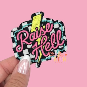 May include: Embroidered patch with the words "Raise Hell" in pink script, outlined in black, on a black and white checkered background. A yellow lightning bolt is behind the text, and the patch is bordered with turquoise beads.