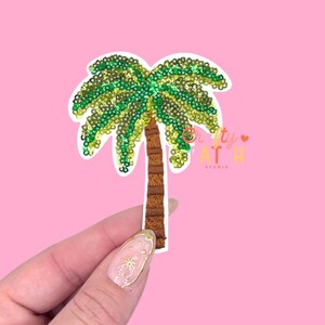 May include: A palm tree patch with green and yellow sequins for the leaves and a brown trunk. The patch is on a white background and is held against a pink background. The text "Crafty AT H Studio" is visible.