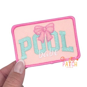 May include: Embroidered patch with a pink background and a pink border. The patch features the words "POOL babe" in turquoise letters, with a pink bow above the "O". The patch is rectangular and has the words "Gritty Patch Studio" in the bottom right corner.