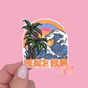 May include: Embroidered patch featuring a beach scene with palm trees, a wave, and a rainbow. The patch has the words "BEACH BUM" in orange letters. The patch is held by a hand against a pink background.