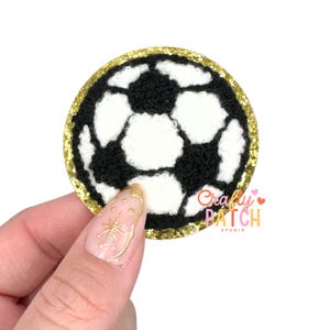 May include: A round, black and white soccer ball patch with a gold glitter border. The patch is held by a hand with a gold nail design. The text "Crafty Patch Studio" is visible.