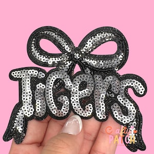 May include: A black sequined patch with the word "Tigers" in silver sequins, topped with a bow. The patch has a black border and is set against a pink background. The patch is held by a hand.