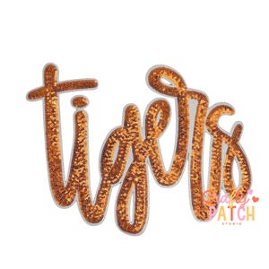 May include: A sequined patch in the shape of the word "Tigers" in a cursive font. The letters are filled with bronze-colored sequins and outlined with a silver border. The patch is on a white background.
