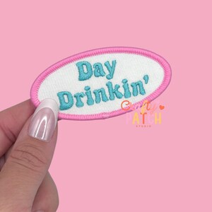 May include: An embroidered patch with the words "Day Drinkin'" in turquoise script on a white oval. The patch has a pink border and is held against a pink background.