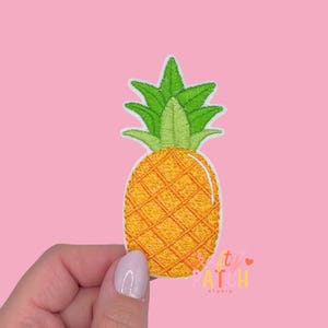 May include: Embroidered pineapple patch with a yellow and orange body and green leaves. The patch has a white border and is held against a pink background. The patch is from the "Pretty Patch Studio".