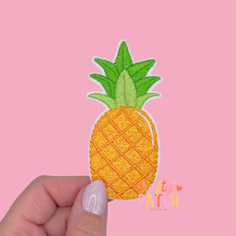May include: Embroidered pineapple patch with a yellow and orange body and green leaves. The patch has a white border and is held against a pink background. The patch is from the "Pretty Patch Studio".