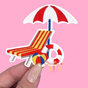 May include: Embroidered patch featuring a beach scene with a red and white striped umbrella, a red and yellow striped beach chair, a life preserver, and a beach ball. The patch has a white border and is set against a pink background.