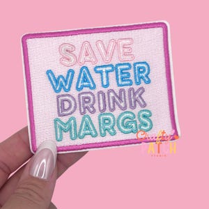 May include: A rectangular embroidered patch with the text "SAVE WATER DRINK MARGS" in pastel colors. The patch has a pink background and a pink border. The words are in a mix of pink, blue, purple, and teal.