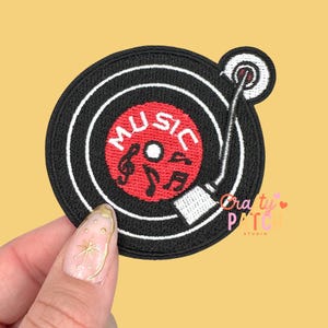 May include: Embroidered patch featuring a black vinyl record design with white concentric circles. The center of the record is red with the word "MUSIC" and musical notes. A white tone arm extends from the record.