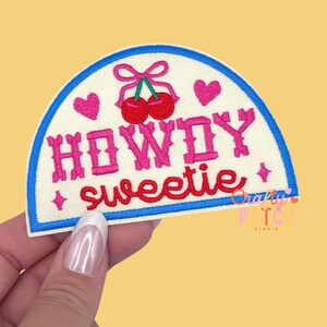 May include: Embroidered patch with a cream-colored background, a blue border, and the words "HOWDY sweetie" in pink. The patch features a pair of red cherries with a pink bow and two pink hearts.