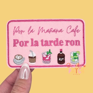 May include: Pink embroidered patch with the text "Por la Mañana Cafe Por la tarde ron." The patch features embroidered images of a coffee cup, iced coffee, cocktail, rum bottle, and a mojito. The patch is rectangular with rounded corners.