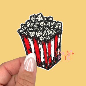 May include: Embroidered popcorn patch with a red and white striped container and overflowing popcorn. The popcorn is white with black outlines. The patch has the text "Crafty Patch Studio".