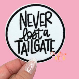 May include: A white circular patch with a black border and the phrase "NEVER lost a TAILGATE" in black script. The patch is held against a pink background.