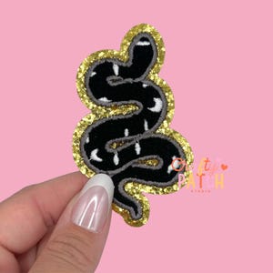 May include: Black snake patch with white accents and a gold glitter border. The snake is embroidered on a black background. The patch is in the shape of a snake and is likely for clothing or accessories.
