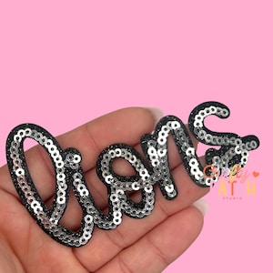 May include: A black sequined patch spelling the word "DREAMS" in a cursive font. The sequins are silver and the border is black. The patch is held in a hand against a pink background.