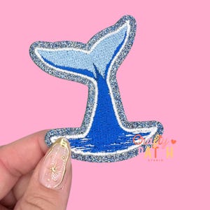 May include: Embroidered patch featuring a blue and light blue whale tail design with a silver glitter border. The patch is on a pink background. The patch has the text "Crafty Patch Studio".