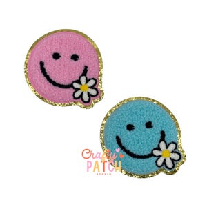 May include: Two embroidered patches featuring smiley faces with a flower in their mouths. One patch is pink, the other is blue. Both have black eyes and smiles, and are outlined with gold glitter.