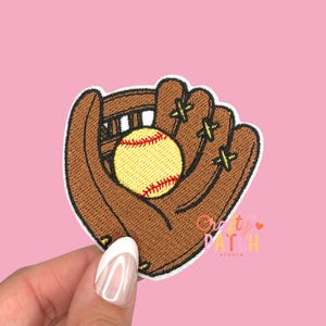 May include: Embroidered patch featuring a softball in a brown glove. The softball is yellow with red stitching. The glove has yellow stitching and is on a white background. The patch is held against a pink background.