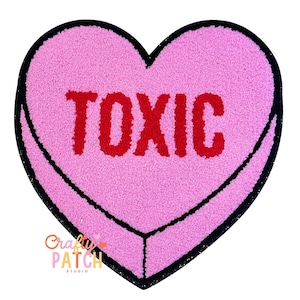 May include: Pink heart-shaped rug with the word "TOXIC" in red letters. The rug has a black border and a textured surface. The rug is a fun and quirky home decor item.