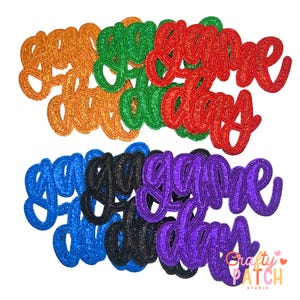 May include: A collection of glittery "Game Day" cutouts in various colors, including orange, green, red, blue, black, and purple. The cutouts are in a cursive font and are likely used for crafting or decorations.