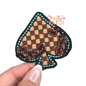 May include: A spade-shaped patch with a brown and tan checkered pattern, adorned with brown floral embroidery. The patch is outlined with a turquoise dotted border and a black edge. The text "Crafty Patch Studio" is visible.