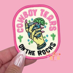 May include: Embroidered patch with the text "COWBOY TEARS ON THE ROCKS" in pink lettering. The patch features a cartoon hand holding a cocktail with a lime wedge. The patch has a pink border and two green cactus designs.