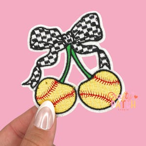 May include: Embroidered patch featuring two yellow cherries designed as softballs, with red stitching and green stems. A black and white checkered bow sits above the cherries. The patch has a white border and is on a pink background.