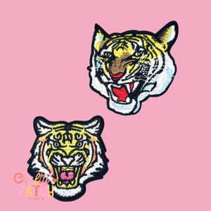 May include: Two embroidered tiger head patches. The tigers have yellow and black stripes, white fur, and open mouths with red tongues. The patches are on a pink background. The text "Cute Lil Patch Studio" is on the bottom left patch.