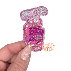 May include: A pink and glittery golf bag patch with golf clubs. The patch has a sparkly, sequined design and the text "Crafty Patch Studio". The golf bag is a vibrant pink color, and the golf clubs are silver.