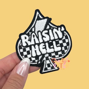 May include: Black and white embroidered patch in the shape of a spade. The patch features the words "RAISIN' HELL" in white, with a checkered pattern in the center. The patch has a black border.