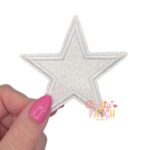 May include: A white, glittery star-shaped patch with a stitched outline. The patch is held by a hand with pink nail polish. The logo "Crafty Patch Studio" is visible in the lower right corner.