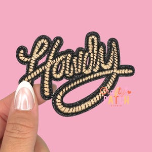 May include: A black and gold embroidered patch with the word "Handy" in a cursive font. The patch has a glittery black border and a light pink background. The patch is held by a person.