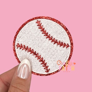 May include: A round baseball patch with a white center and red stitching. The border is red glitter. The patch is held by a hand with a pink background. The text "Crafty Patch Studio" is visible.