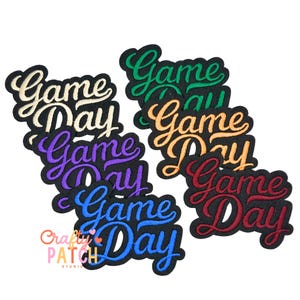 May include: A collection of embroidered patches with the words "Game Day" in a cursive font. The patches feature various color combinations, including white, green, orange, red, purple, and blue, all on a black background. The "Crafty Patch Studio" logo is also visible.