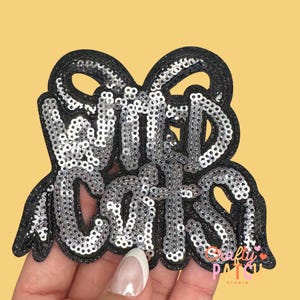 May include: A black sequined patch with the words "Wild Cats" in silver sequins. The patch has a bow-like shape at the top and a scalloped edge. The background is a solid yellow.
