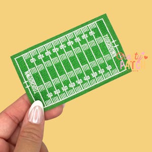 May include: Green and white embroidered patch depicting a football field with yard lines and the word "TOUCHDOWN". The patch is rectangular and held by a hand against a yellow background. The patch has the text "TOUCHDOWN" and numbers indicating yard lines.