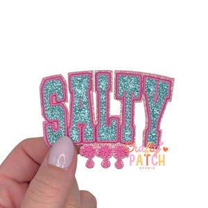 May include: A pink and teal glitter patch with the word "SALTY" in a collegiate font. The patch has a pink border and teal glitter filling. Below the word are three pink palm trees. The patch is from Crafty Patch Studio.