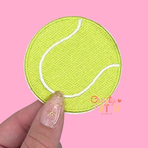 May include: A round, embroidered patch in the shape of a tennis ball. The patch is primarily a bright yellow-green color with white curved lines and a white border. The patch is held against a pink background.